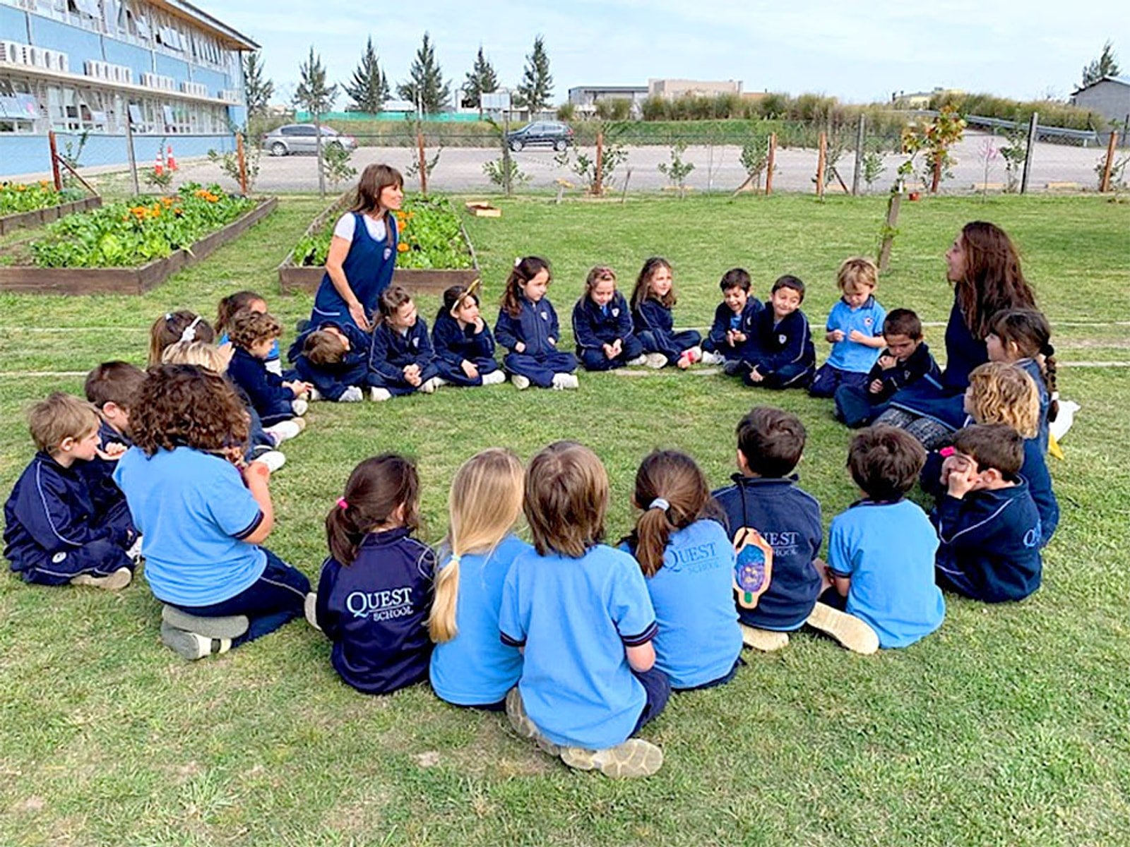Ronda de alumnos de inicial sentados en el césped junto a la huerta con sus maestras